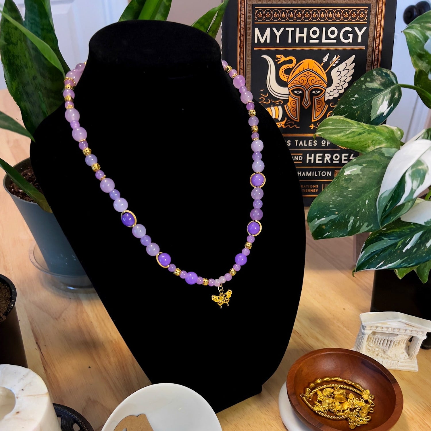 A short length necklace, with  golden butterfly charm at its center, comprised of purple beads in varying shades and golden accent pieces. The necklaces is on a black stand with a book entitled 'Mythology' in the background.
